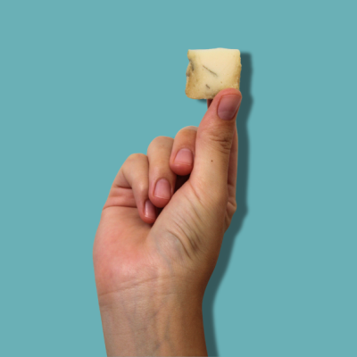 Mão a segurar pequeno cubo de queijo amarelo com manchas escuras sobre fundo azul.