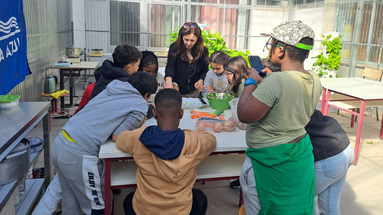 Crianças e mulher numa cozinha em torno de uma mesa com legumes.