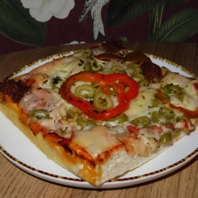 Fatia quadrada de pizza com queijo, azeitonas, pimentos e linguiça num prato branco sobre mesa de madeira