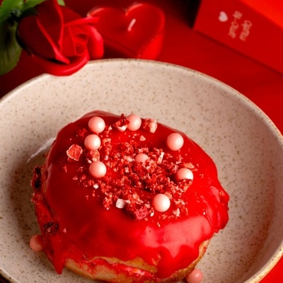 Donut com glacê vermelho e decoração de pérolas e confeitos vermelhos num prato bege, com rosa e vela em forma de coração em fundo vermelho.