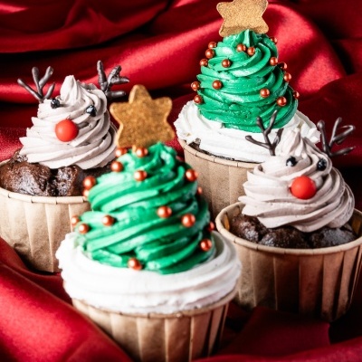 Cupcakes decorados de Natal com creme verde, branco e castanho, estrelas douradas e decoração de rena, sobre tecido vermelho.