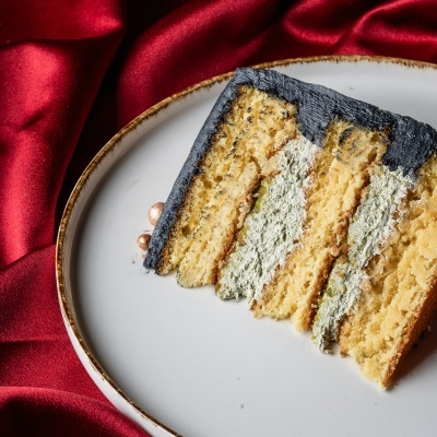 Fatia de bolo com camadas amarelas e cobertura preta num prato branco sobre tecido vermelho