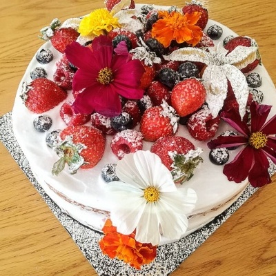 Bolo redondo decorado com frutos vermelhos e flores comestíveis em cima, com cobertura branca, sobre base preta com pontos brancos e mesa de madeira.