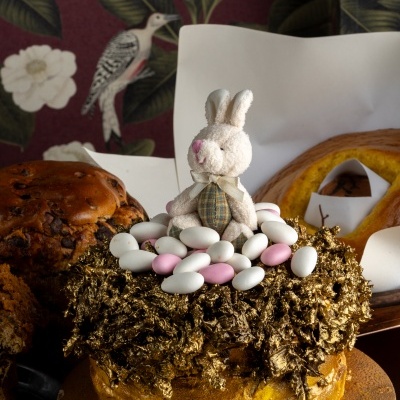 Bolo decorado com cobertura dourada, amêndoas açucaradas e coelhinho de peluche, em ambiente com papel de parede floral