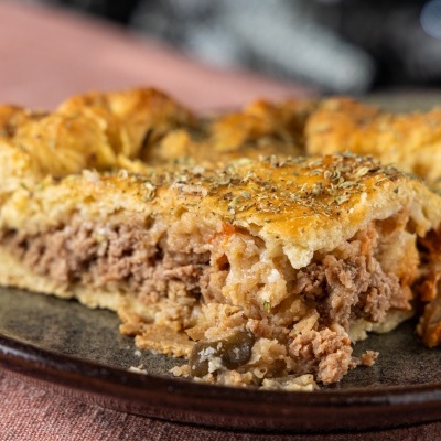 Fatias de empadão de carne com massa dourada e ervas num prato castanho sobre toalha rosa