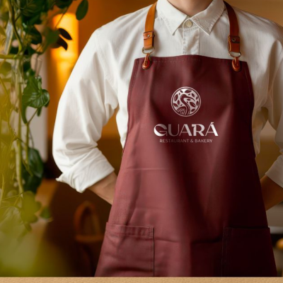 Avental vinho com logótipo e texto branco, alças de couro castanho, usado com camisa branca.