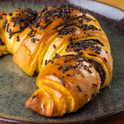 Croissant com granulado de chocolate num prato cerâmico azul escuro sobre mesa de madeira