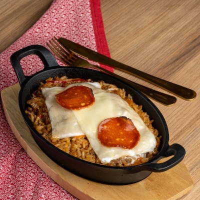 Prato de ferro fundido com arroz, queijo e pepperoni sobre base de madeira e toalha vermelha
