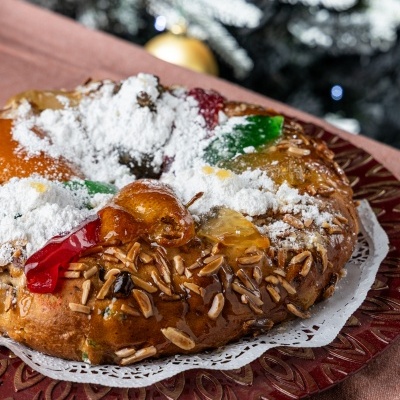 Bolo rei com frutas cristalizadas, amêndoas e açúcar em pó num prato vermelho, com árvore de Natal ao fundo.