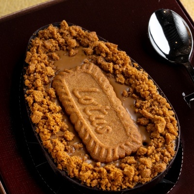 Ovo de chocolate com creme caramelo, biscoito Lotus e granulado caramelo num prato preto com colher.