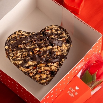 Bolo de chocolate em forma de coração com crumble, dentro de caixa vermelha com texto e vela em forma de coração ao fundo