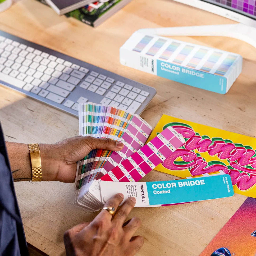 *NOVO* Color Bridge Guide Set COATED & UNCOATED Amostras de cores Pantone em leque na mão sobre secretária de madeira com teclado e caixas coloridas