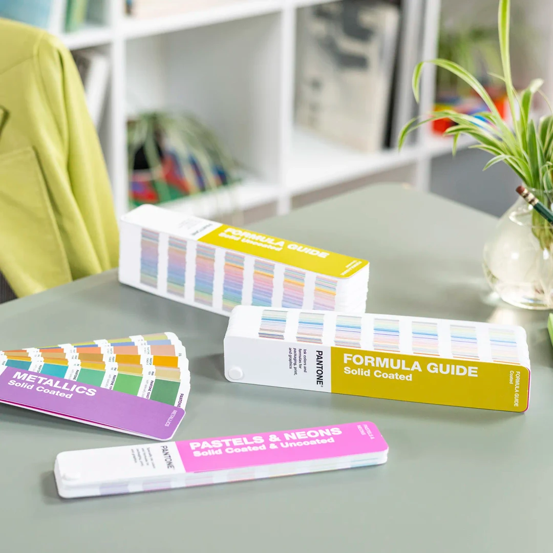 Guias de cores Pantone variando entre sólidos, metálicos e neons sobre uma mesa verde.