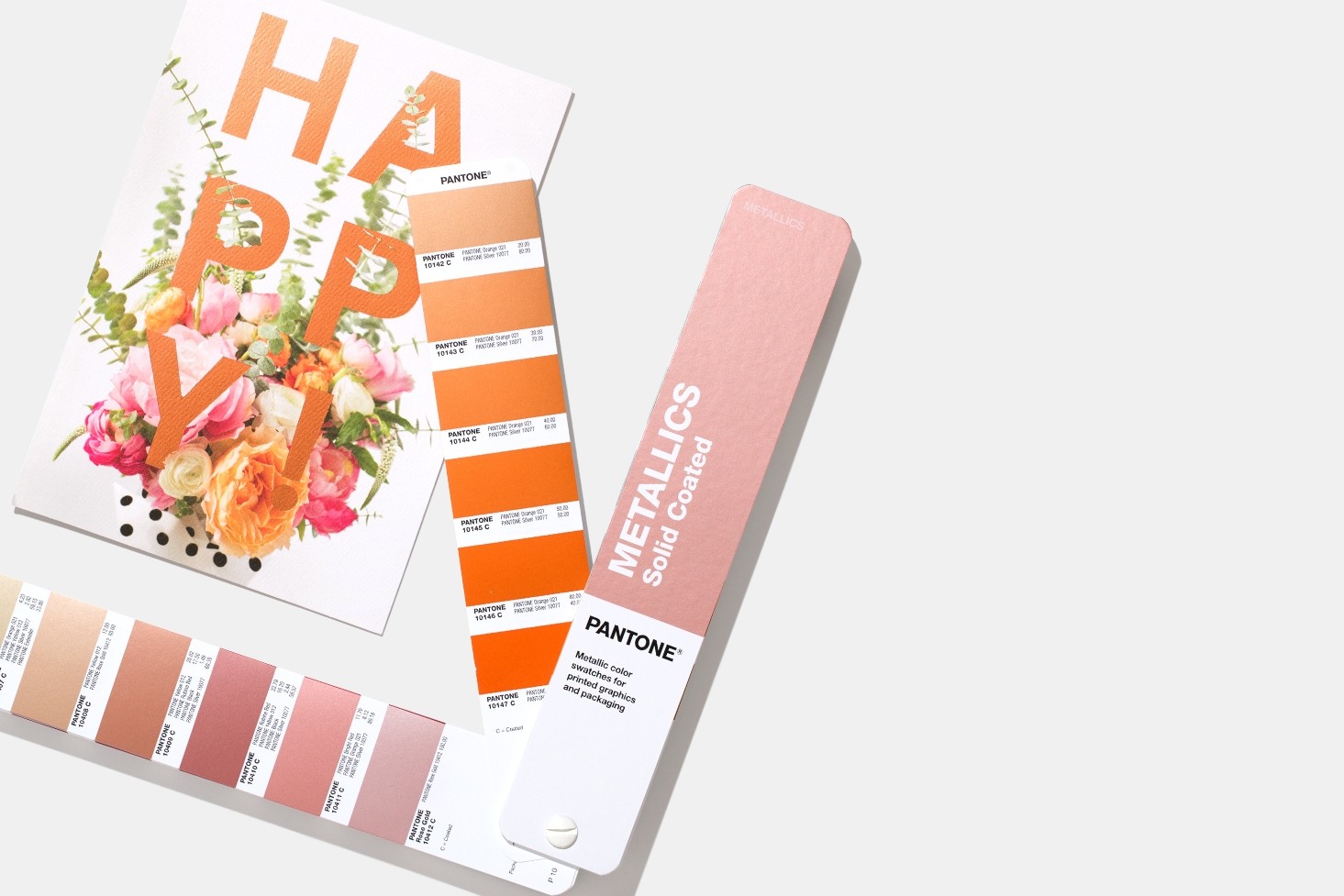 Metallics Guide Cartões Pantone com amostras metálicas em tons laranja e rosa ao lado de cartão com texto HAPPY e flores