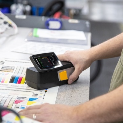 Dispositivo portátil preto com ecrã em cima de folhas de amostras coloridas numa mesa de trabalho.