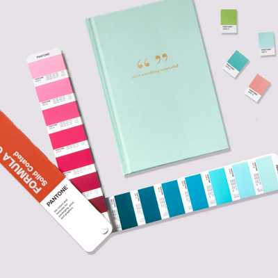 Caderno azul claro com texto dourado e amostras Pantone em rosa, azul e verde