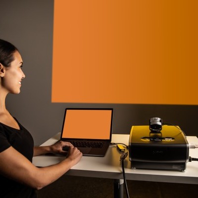 Mulher com computador portátil e projetor numa mesa, projetando imagem laranja numa parede cinzento-escura