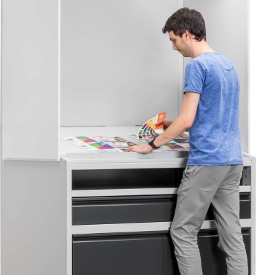 Mesa de trabalho branca com painel de luz e homem a analisar amostras de impressão