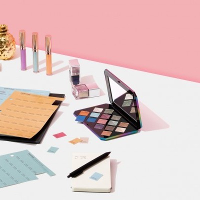 Paleta de sombras, batons, caneta, caderno e fichário sobre mesa branca com fundo rosa