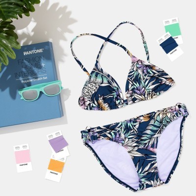 Biquíni floral azul e lilás com alças cruzadas, livro Pantone, paletas de cores e óculos de sol verdes.