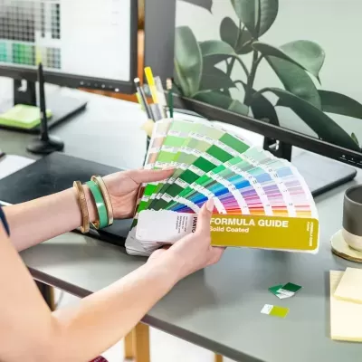 Cartela de cores Pantone FORMULA GUIDE Solid Coated nas mãos com fundo de escritório com computador e acessórios.