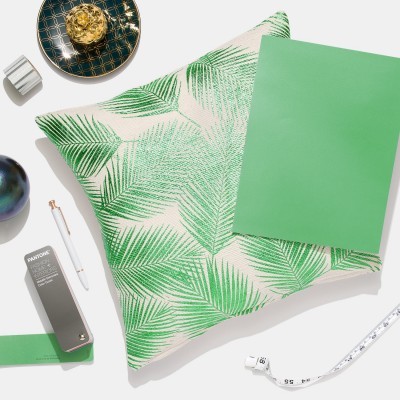 Almofada branca com folhas verdes estampadas rodeada por objetos diversos sobre fundo branco