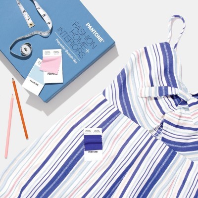 Vestido às riscas azuis, lilases e laranja ao lado de fita métrica, lápis, amostras Pantone e livro azul