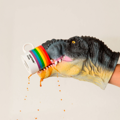 Luva de dinossauro a segurar caneca branca com faixas coloridas PANTONE a escorrer líquido laranja