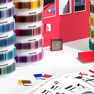 Conjuntos de amostras de cores Pantone em discos rotativos transparentes sobre mesa branca com relógio, livros vermelhos e catálogo de móveis.