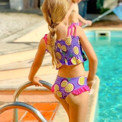 Biquíni infantil cor roxa com padrão de maracujá e detalhes rosa junto à piscina