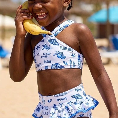 Menina na praia com fato de banho branco e azul, segurando uma banana junto à orelha