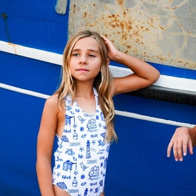 Menina loira com fato de banho branco estampado com motivos marítimos azuis, encostada a um barco azul e branco com sinais de ferrugem.