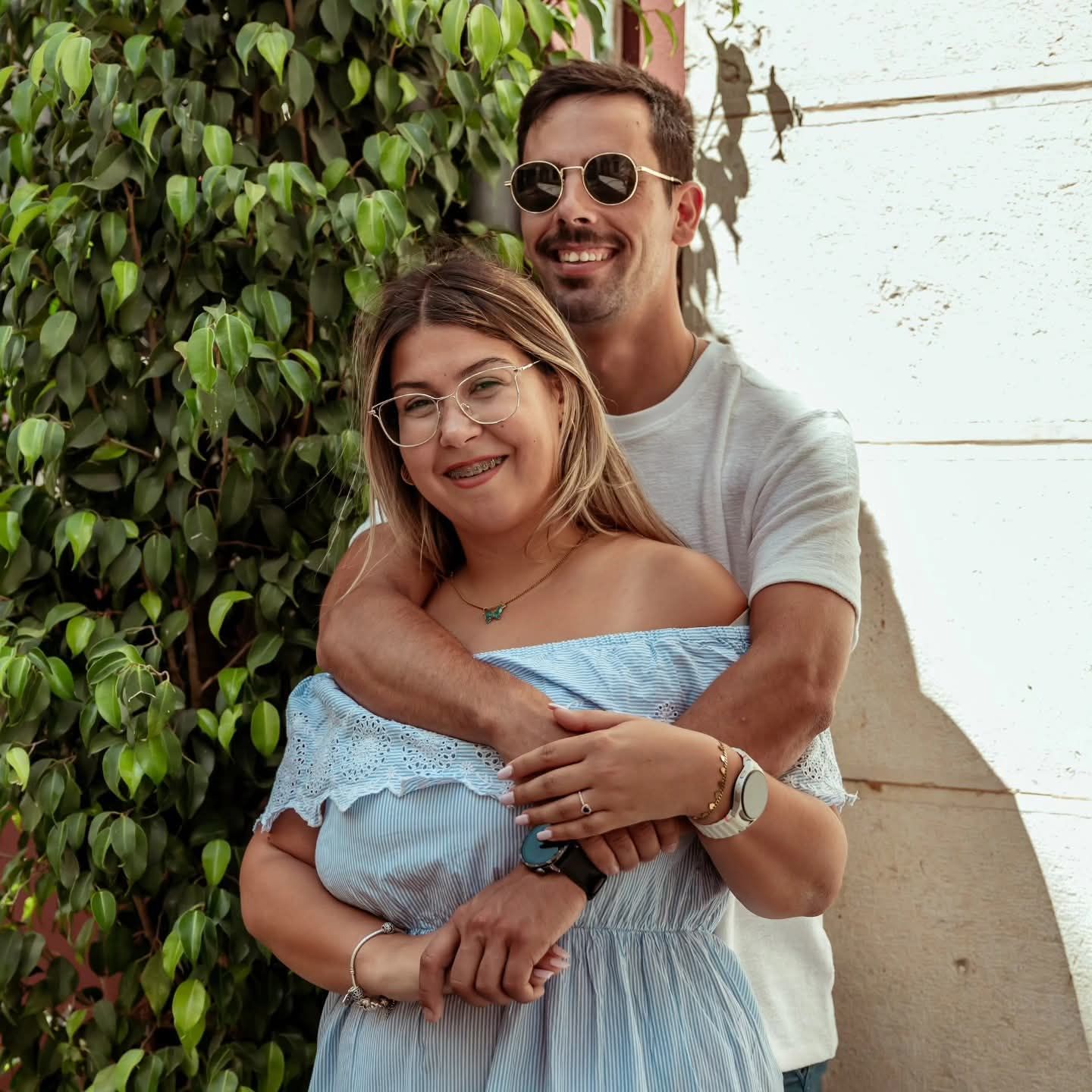 Casal jovem abraçado com roupa casual e acessórios, em ambiente exterior com parede e plantas