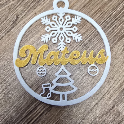 Enfeite de Natal redondo com nome Mateus em letras douradas e elementos brancos sobre madeira.