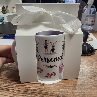Caneca branca com ilustrações de treino e texto Personal Trainer dentro de caixa branca com fita branca.