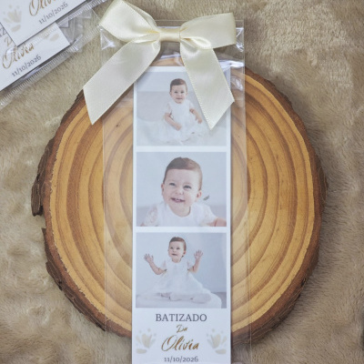 Marcador de página com fotos de bebé e texto de batizado, com laço de cetim creme, sobre madeira.