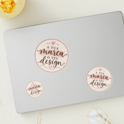 Laptop prateado com autocolantes redondos personalizados com texto em português e itens de escritório em fundo branco.