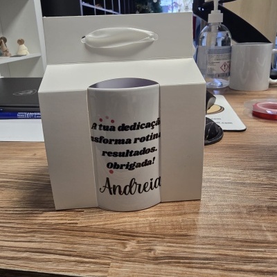Caneca branca com texto de agradecimento dentro de caixa branca com pega sobre mesa de madeira.