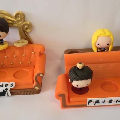 Sets de decoração com sofás laranja e bonecos pequenos, um com rato amarelo no cabelo, e placas com o texto FRIENDS.