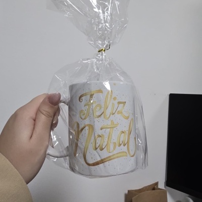 Caneca branca com texto dourado Feliz Natal embrulhada em plástico transparente