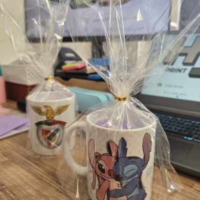 Canecas de cerâmica embaladas em plástico transparente com ilustrações distintas, sobre mesa de madeira.