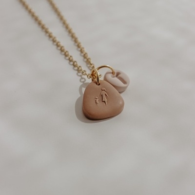 Colar dourado com dois pendentes em argila castanha e clara, sobre fundo branco.