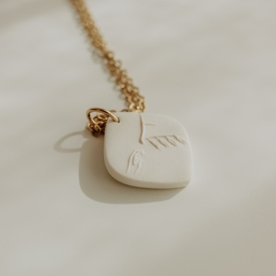 Colar dourado com pendente bege com relevo de rosto e mão.
