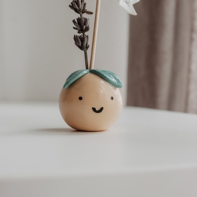 Vaso cerâmico decorativo redondo com carinha sorridente, folhas verdes, flor seca e bandeira pequena branca