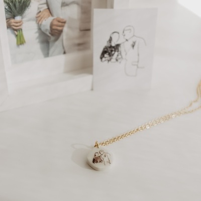 Colar com pingente redondo branco com desenho de casal, fotografia e cartão com ilustração na mesa branca