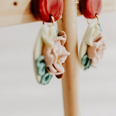 Brincos pendurados num suporte de madeira clara, com base vermelha e decoração colorida em tons pastel