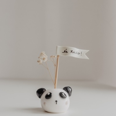 Vaso de cerâmica em forma de panda com flores secas e bandeira com texto em português.