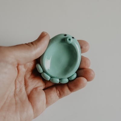 Objeto cerâmico verde claro em forma oval com detalhes de patas e olhos pretos