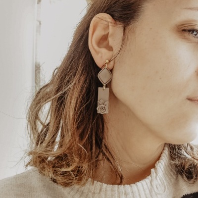 Brinco pendente bege com detalhe de flor usado por pessoa com suéter bege e riscas pretas.