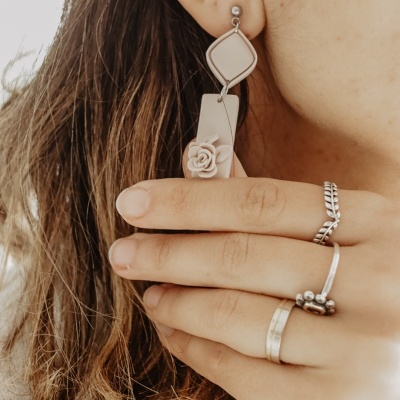 Brinco pendente bege com flor e vários anéis em mão feminina